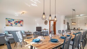 Un espace repas moderne avec une table en bois dressée pour six personnes, entourée de chaises. Un grand tableau abstrait orne le mur, ainsi qu’une horloge et un téléviseur posé sur un meuble.
