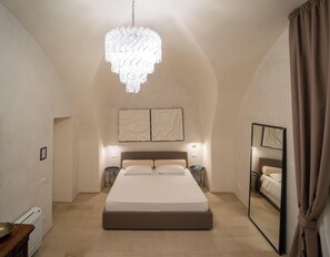 Un dormitorio con una cama grande, una araña, dos lámparas de pared, un espejo y una puerta que da a otra habitación.