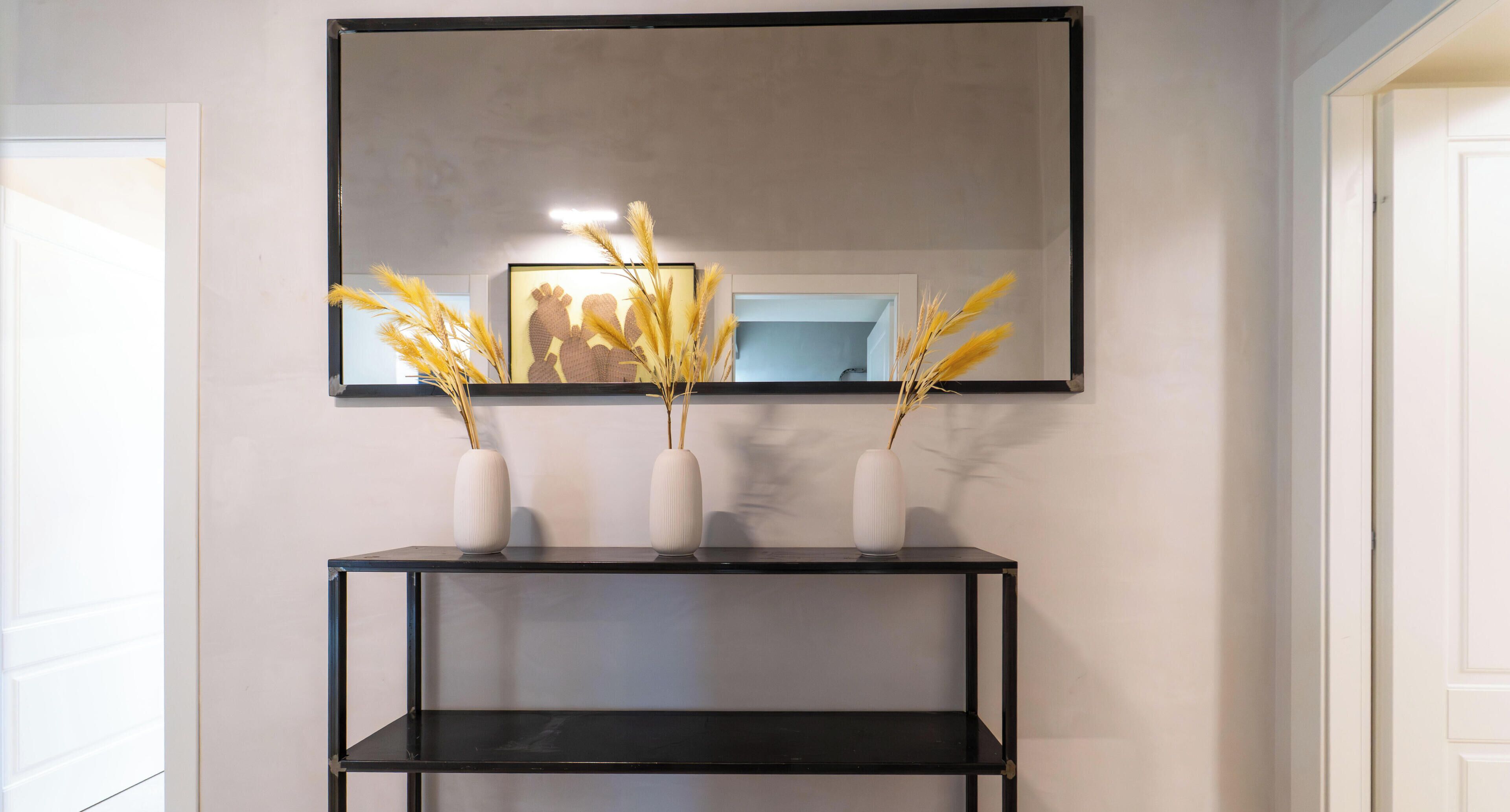 A wall-mounted mirror above a black console table with three white vases containing dried plants.