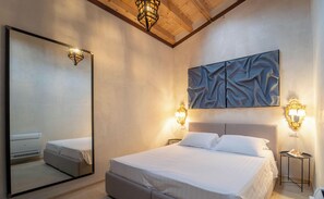 A bedroom with a large mirror, a bed with white bedding, two bedside tables with lamps, and a ceiling with exposed beams.