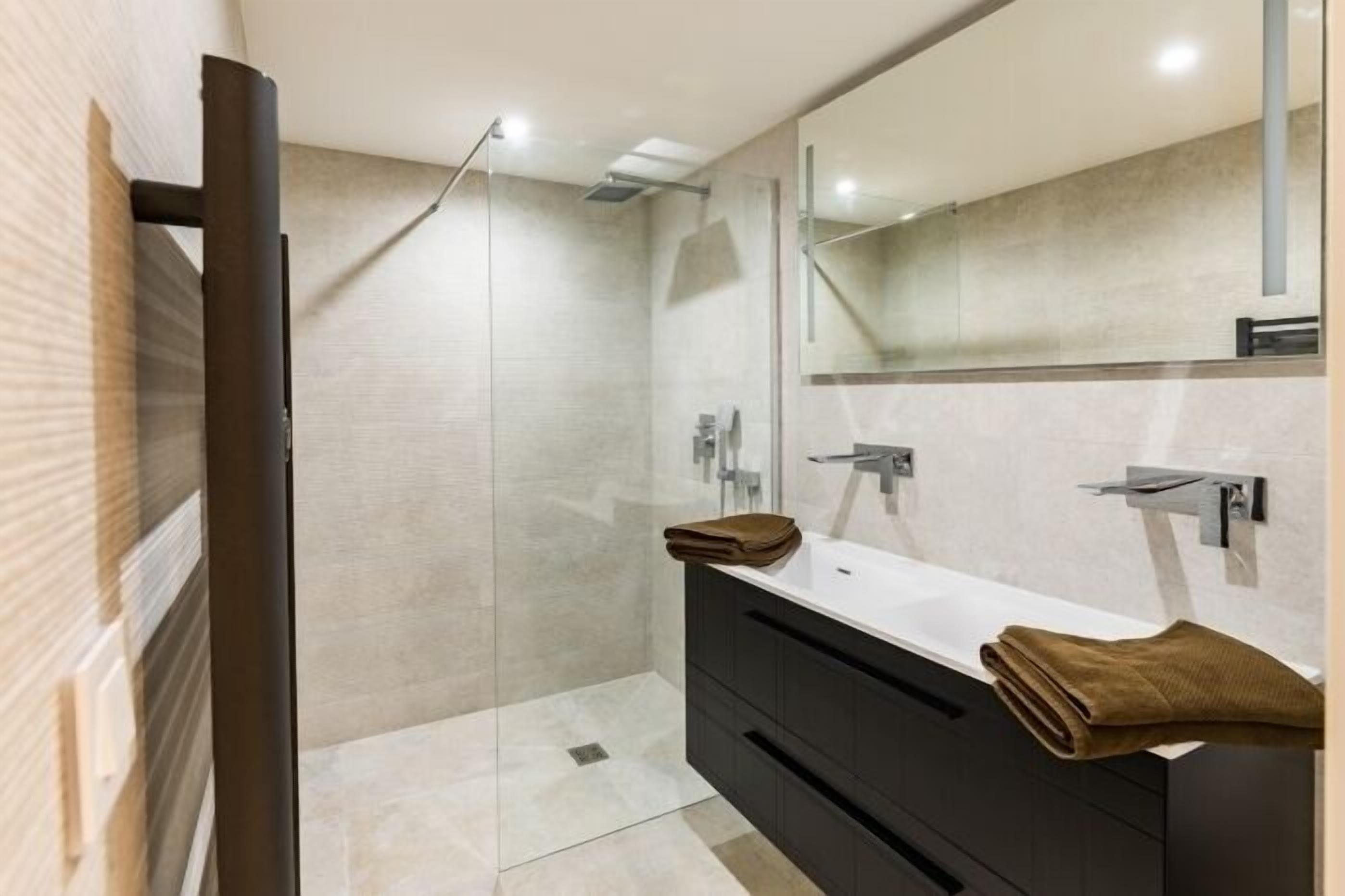 A modern bathroom with a glass-enclosed shower, a white sink, dark cabinets, and a large mirror.