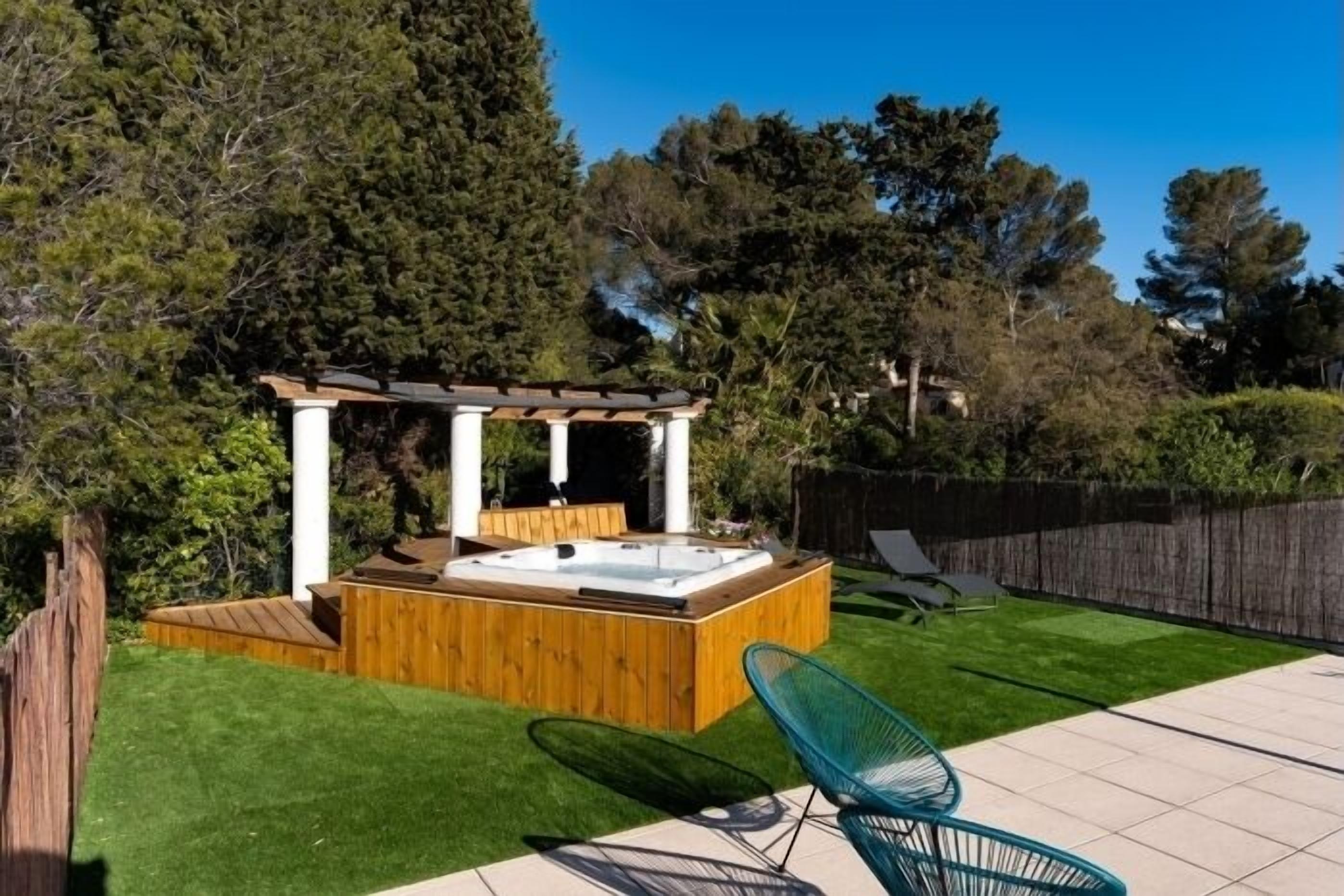 Un jacuzzi con cubierta de madera, rodeado por una pérgola de madera, en un patio trasero con césped y valla de madera.