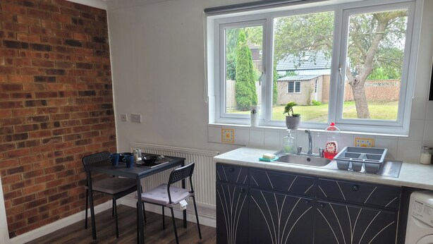 A modern kitchen with a brick wall, a dining table with chairs, a window with a view of greenery, and a built-in washing machine. - Charming Luton Apartment Retreat (Luton)