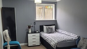 A bedroom with a bed, a desk, a chair, a standing fan, and a window. - Charming Luton Apartment Retreat (Luton)