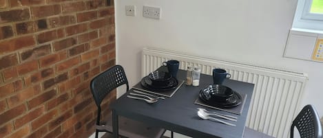 A dining area with a brick wall, a table set with black plates and silverware, two chairs, and a window with blinds.