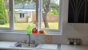 A kitchen with a stainless steel sink, a window with a view of a house and garden, and a dishwasher. - Charming Luton Apartment Retreat (Luton)