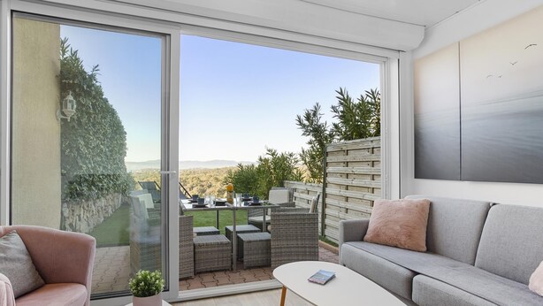 A modern living room with a grey sofa, a white coffee table, and a pink armchair. A large sliding glass door leads to an outdoor patio with a view of the countryside.