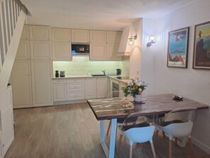 Una cocina compacta con armarios de colores claros, un microondas y un lavabo. Hay una mesa de comedor con sillas y un florero con flores.
