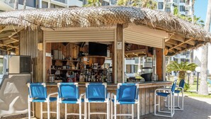 Un bar junto a la playa con techo de paja, paredes de madera y una barra con varias botellas y un televisor. Hay cuatro sillas azules y una mesa frente al bar.