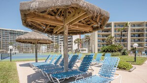 Zona de piscina con sillones azules y sombrillas de paja, rodeada por un edificio de hotel de varios pisos.