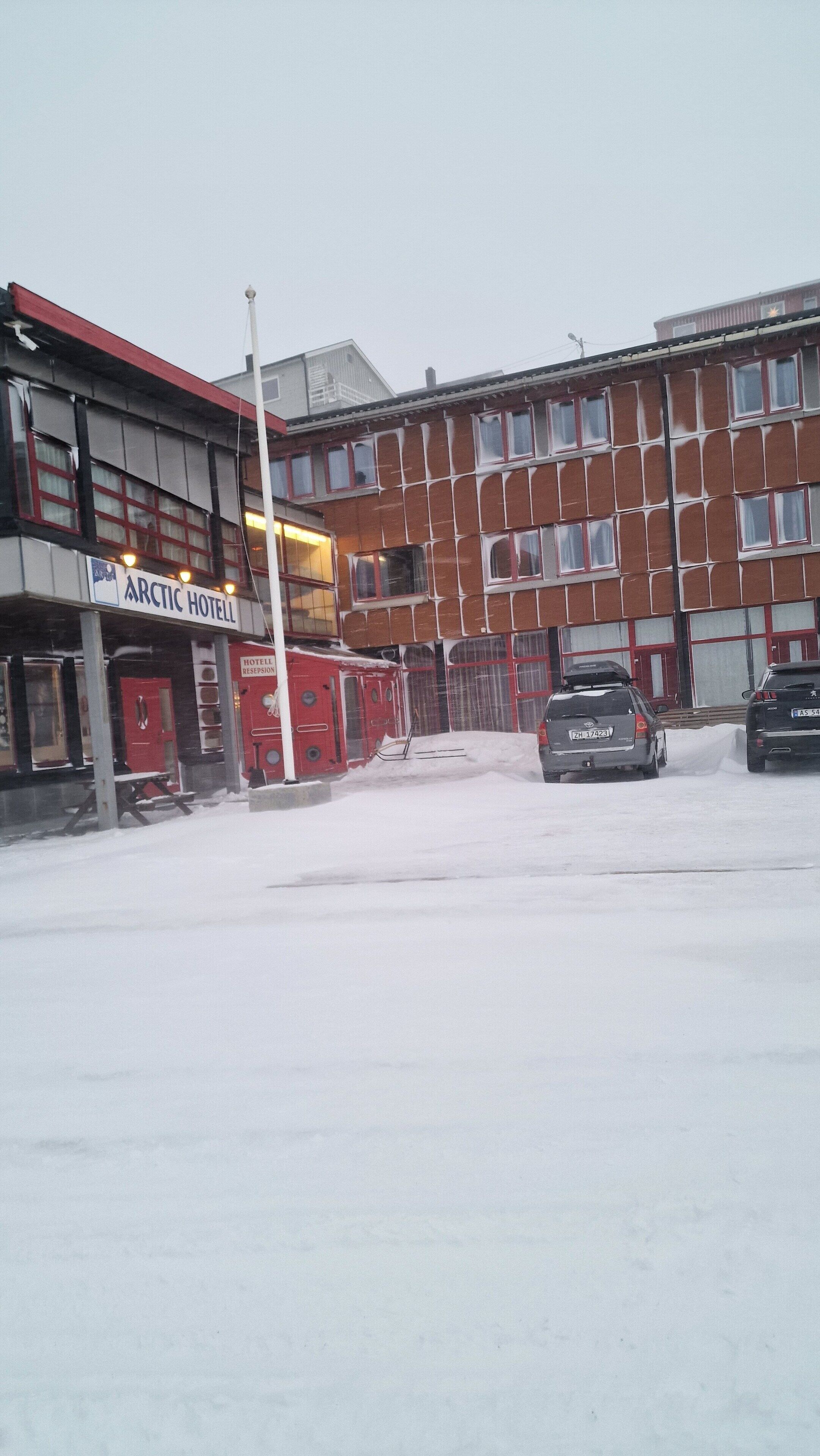 Un hotel con fachada roja y blanca, un letrero que dice "Arctic Hotel" y un estacionamiento cubierto de nieve con dos coches.