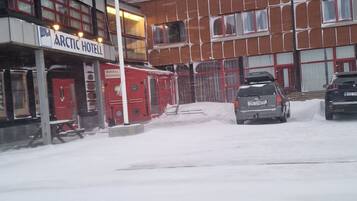 Un hotel con fachada roja y blanca, un letrero que dice "Arctic Hotel" y un estacionamiento cubierto de nieve con dos coches.