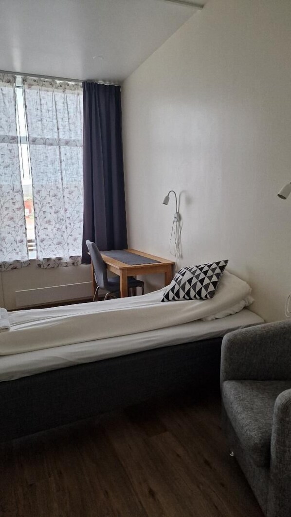 A small, single-bed room with a desk and chair, a window with curtains, and a bedside lamp. - Mehamn Arctic Hotell AS (Gamvik)