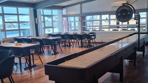 A spacious indoor area with long tables and chairs, large windows, and a ceiling with visible beams and lighting fixtures. - Mehamn Arctic Hotell AS (Gamvik)