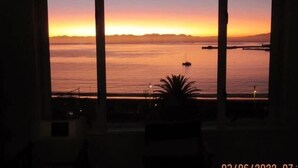 A room with a large window overlooking a calm sea at sunset. - Simon’s Town Seaside Apt | Views, Balcony, Parking (Cape Town)