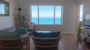 A living room with a view of the sea, a bookshelf, a television, and wicker furniture. - Simon’s Town Seaside Apt | Views, Balcony, Parking (Cape Town)