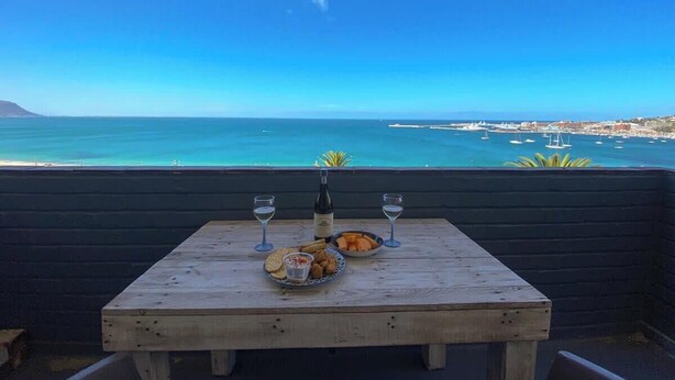 A wooden table with a bottle of wine, two glasses, and a plate of food on a balcony overlooking a coastal view with boats and buildings. - Simon’s Town Seaside Apt | Views, Balcony, Parking (Cape Town)
