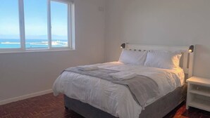 A bedroom with a large window, a bed, and a nightstand. - Simon’s Town Seaside Apt | Views, Balcony, Parking (Cape Town)