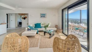 Una sala de estar moderna con sofá, una mesa de centro y sillas de mimbre, un balcón con mobiliario exterior y vistas a la ciudad.