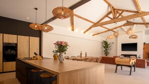 Eine moderne Küche mit einer zentralen Kochinsel, Barhocker und einem Essbereich inklusive Sofa und Fernseher.