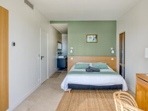 Quarto com cama, mesa de cabeceira, cadeira, banheiro visível por uma porta aberta e um quadro na parede.