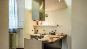 Una cocina moderna con una isla central, estufa integrada y microondas. Se aprecia una ventana con cortinas translúcidas, un radiador blanco y un reloj en la pared.