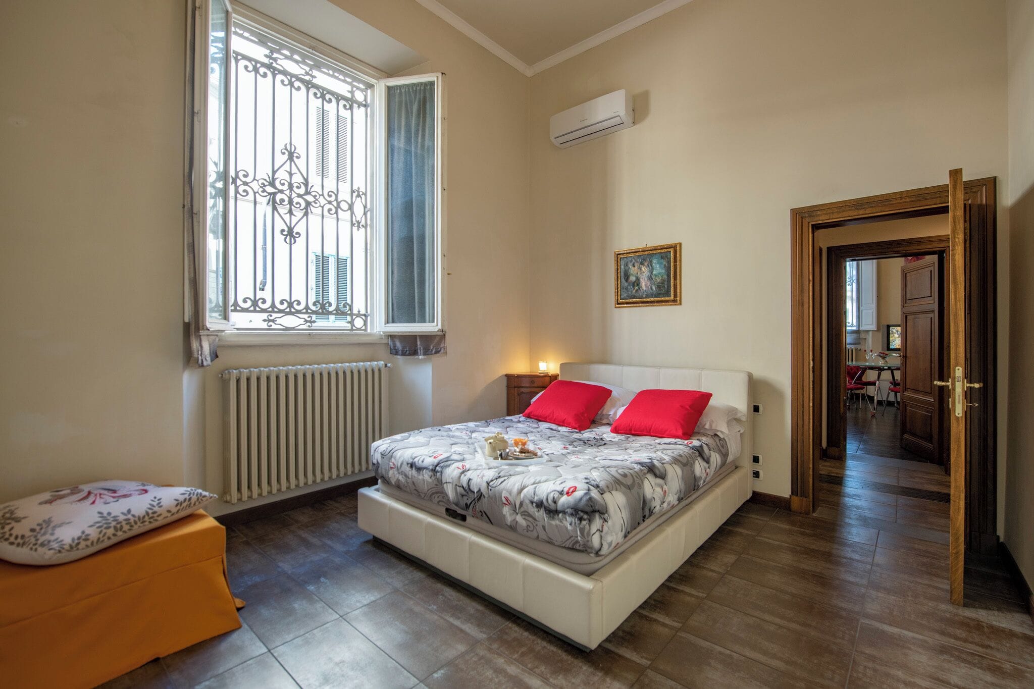A bedroom with a bed, a radiator, a window with bars, a bedside table, a nightstand, and a door leading to another room.