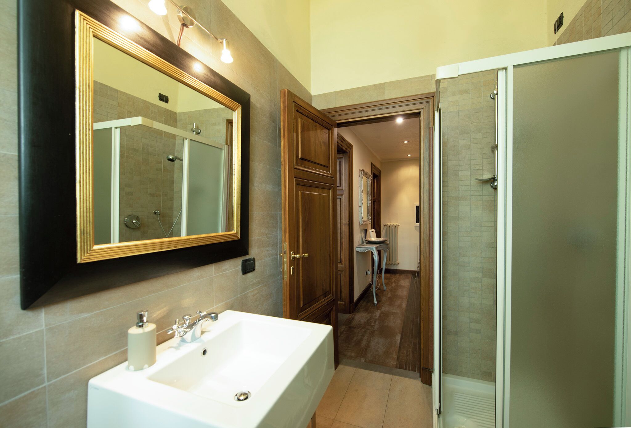 Une salle de bain avec un grand miroir, un lavabo, une douche et une porte donnant sur une autre pièce.