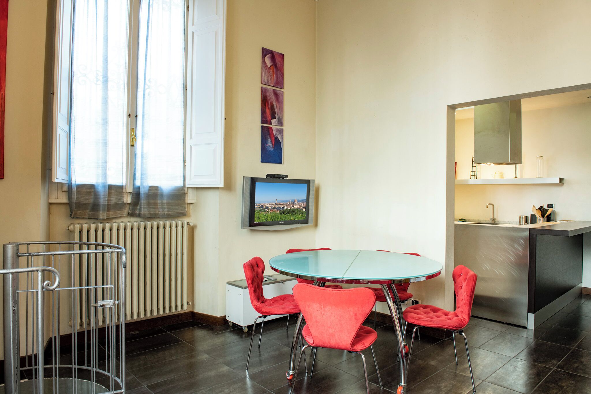 A modern dining area with a round table and red chairs, a television mounted on the wall, and a kitchen area with a stainless steel refrigerator.