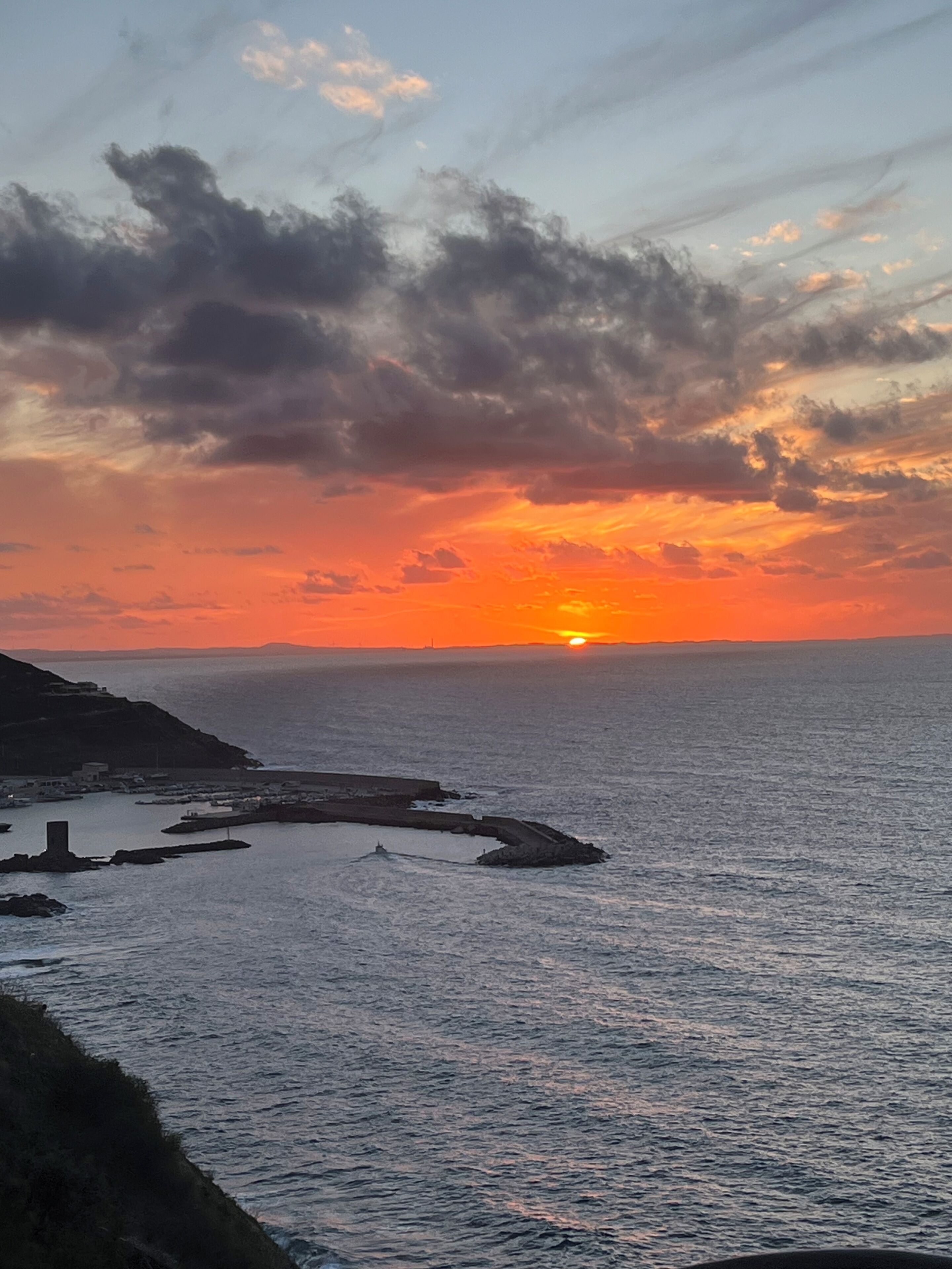 Un coucher de soleil sur la côte, avec une mer calme, une rive rocheuse et des bâtiments situés au loin.