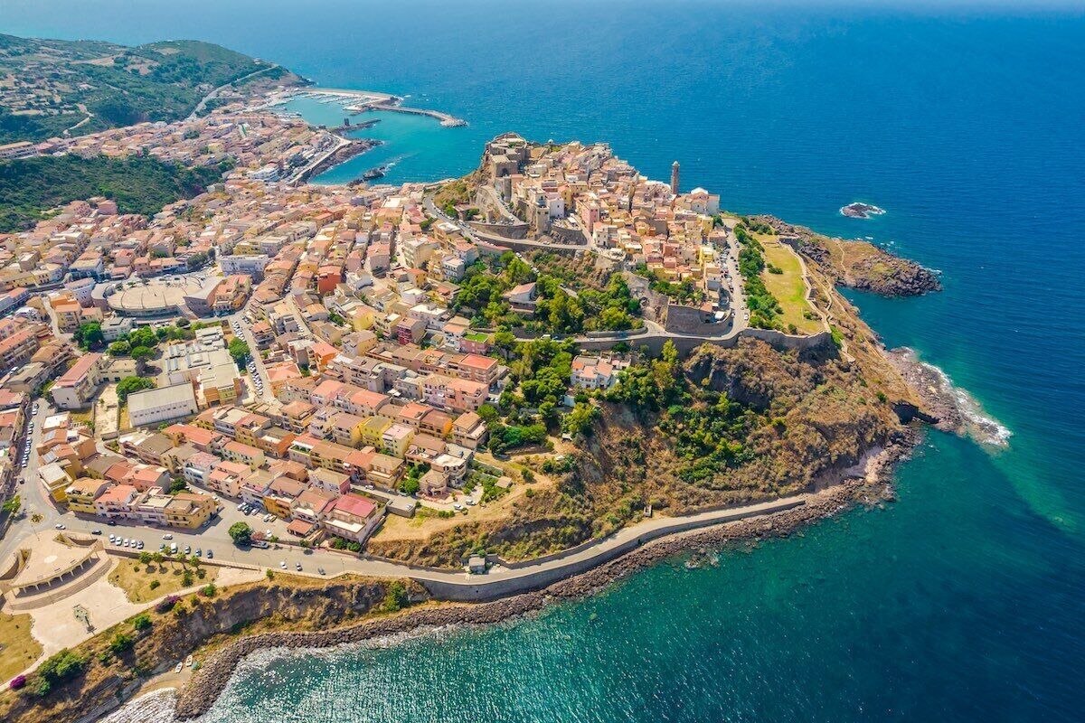 Vista aerea di una cittadina costiera con un’area fortificata centrale, edifici residenziali circostanti e una chiara visuale della costa.