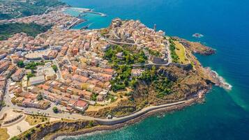 Vista aerea di una cittadina costiera con un’area fortificata centrale, edifici residenziali circostanti e una chiara visuale della costa.