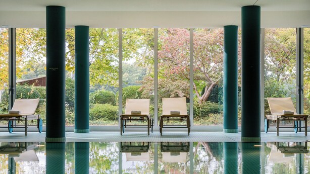 Uma área de piscina interior tranquila, com três espreguiçadeiras, janelas amplas e o reflexo de árvores.