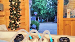 Una baita in legno con una vasca idromassaggio, un muro di legno con una pianta rampicante e vista su una strada con auto e alberi.