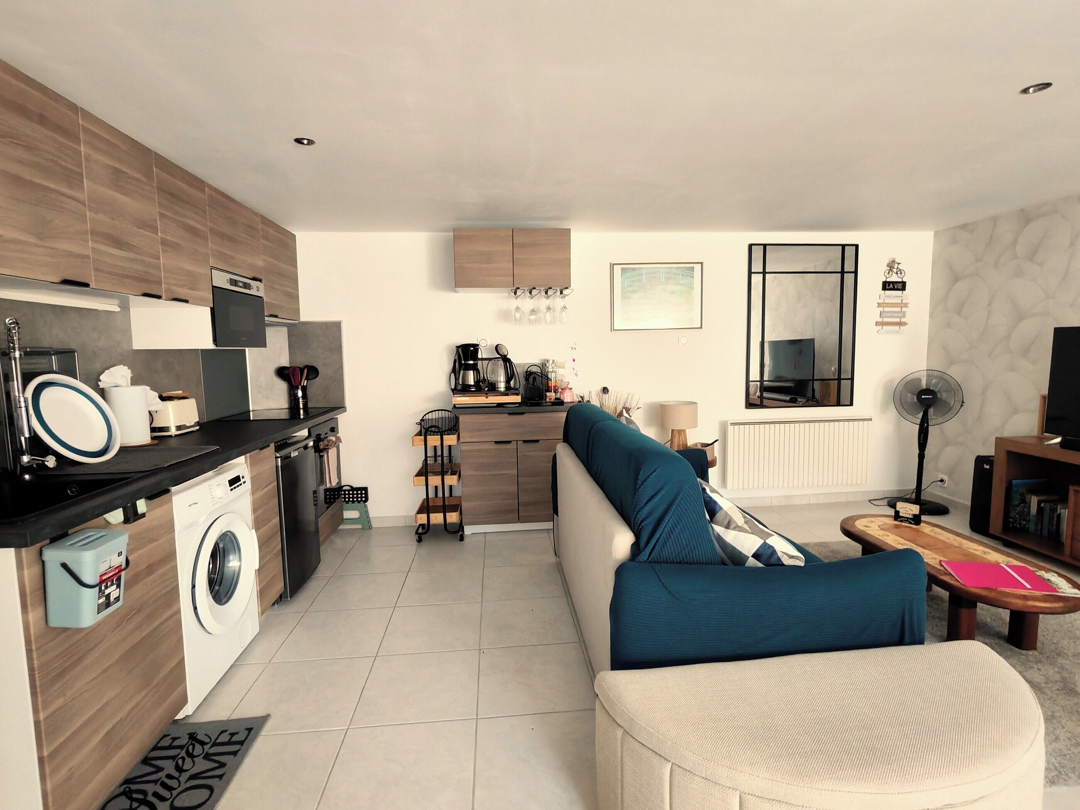 Une cuisine moderne équipée d’un lave-linge intégré, un coin repas avec un canapé et une table, et un salon avec une télévision et un ventilateur.