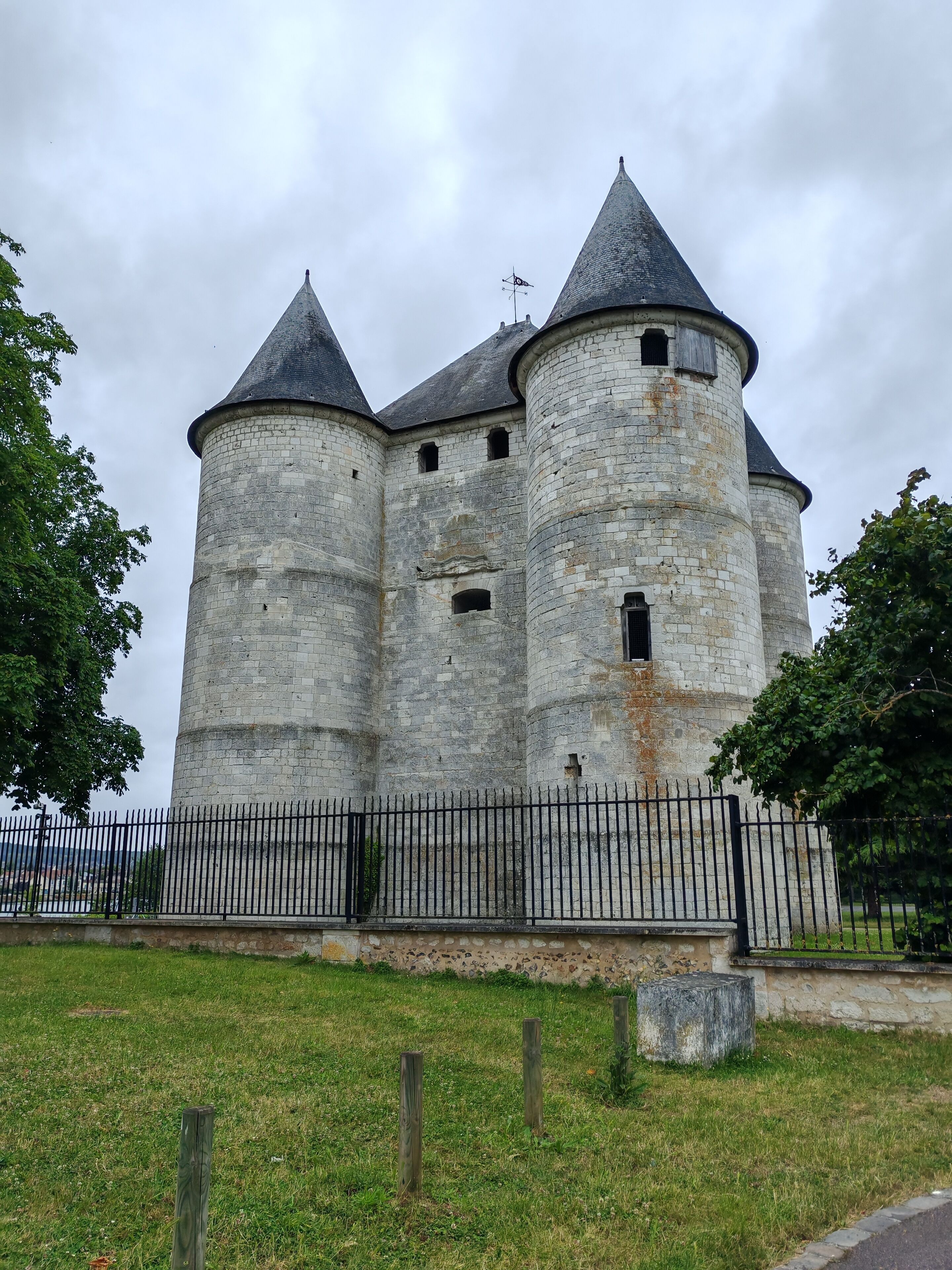 Un château de pierre historique, doté de trois tours, avec une clôture en métal noir au premier plan et une pelouse devant.