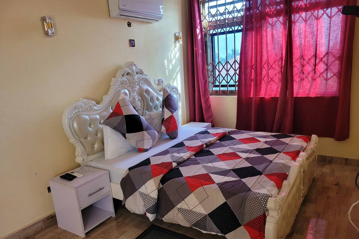 Una cama bien hecha con un edredón estampado, una mesita de noche blanca con una lámpara y una ventana con cortinas rojas.