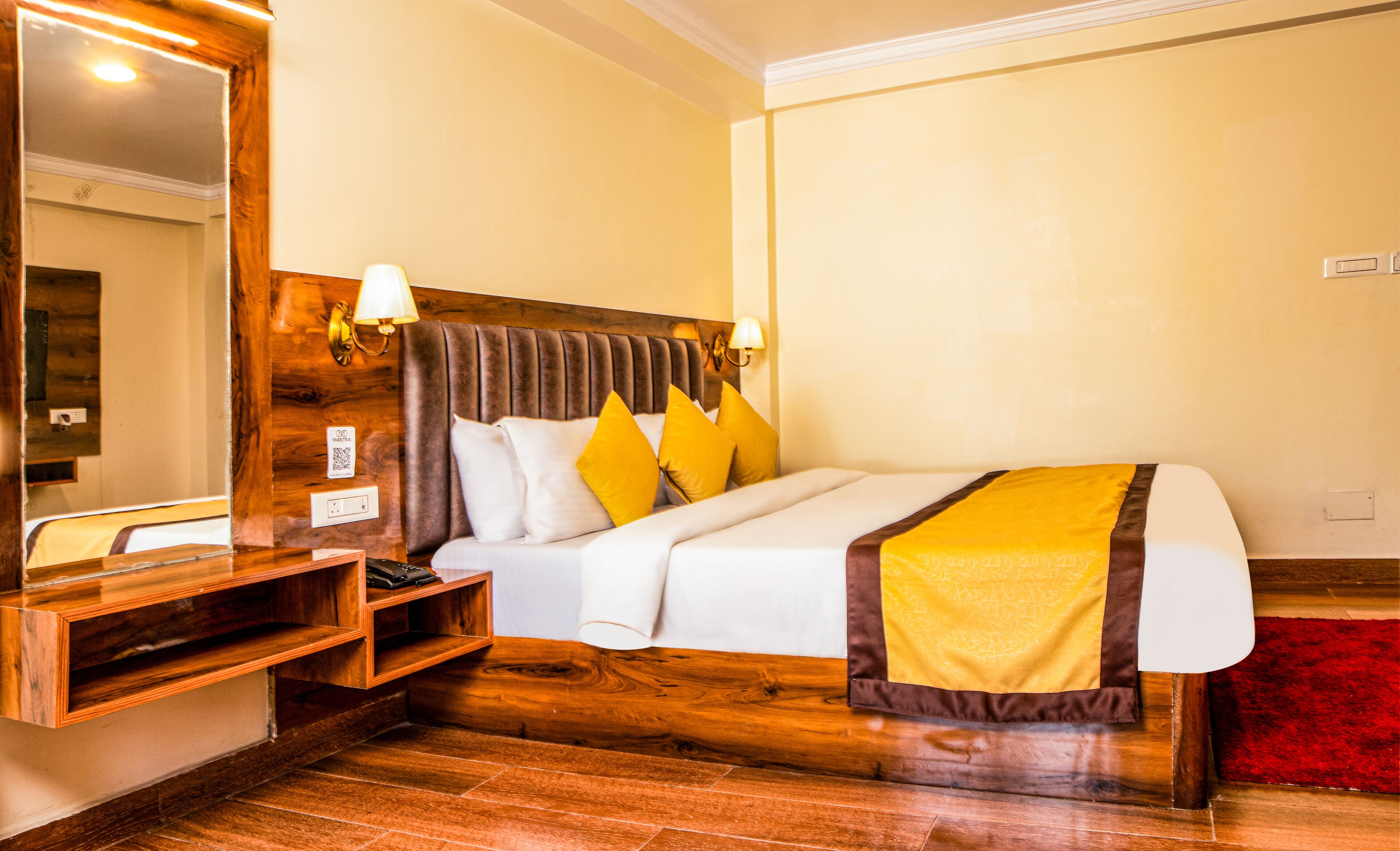 A hotel room with a wooden headboard, a bed with white and yellow bedding, a nightstand with a lamp, and a red carpet.