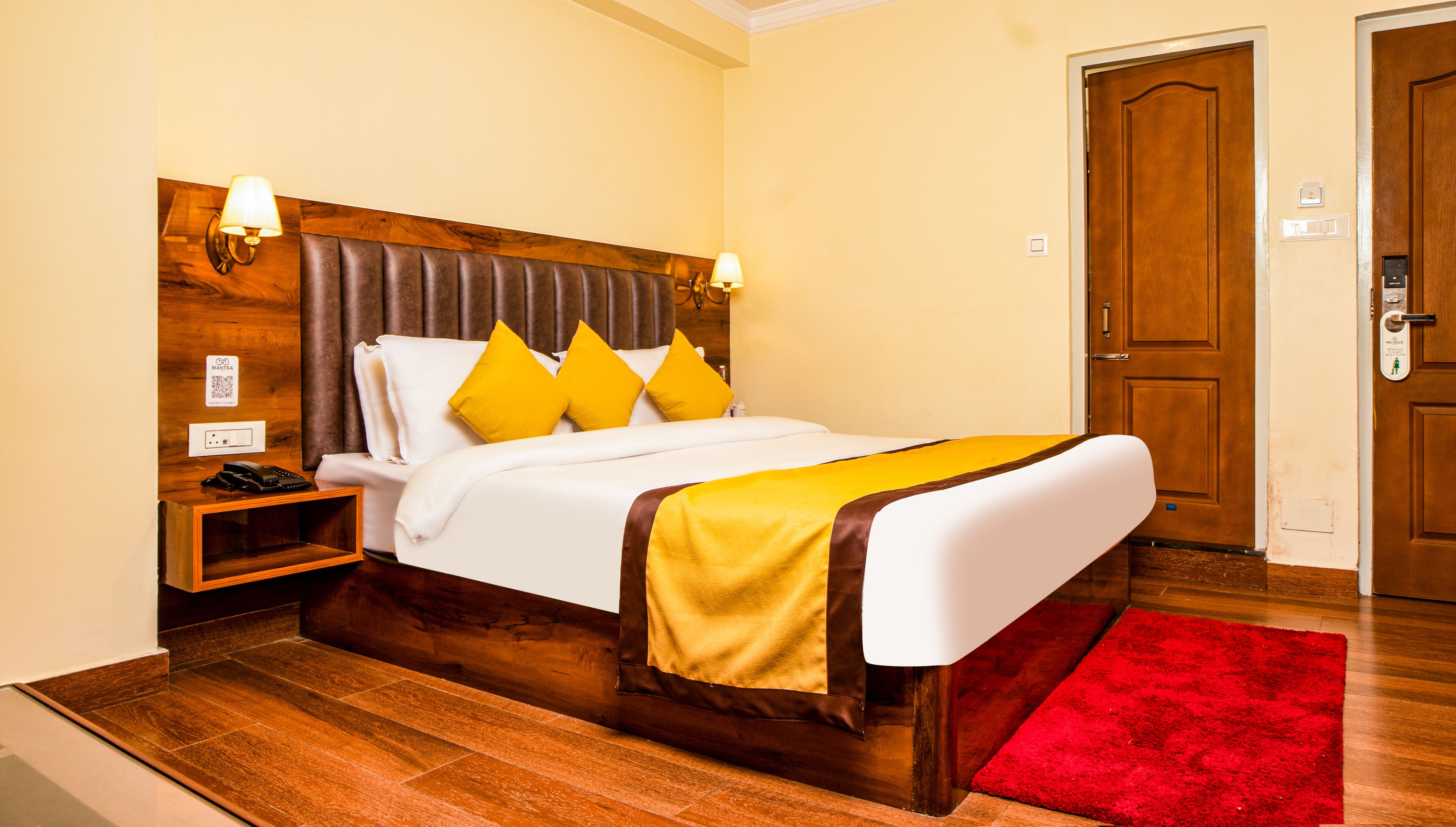 Ein Hotelzimmer mit einem hölzernen Kopfende, einem weißen Bett mit gelben Kissen, einem roten Teppich und einem Nachttisch aus Holz mit einem Telefon.