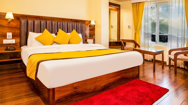 A hotel room with a wooden headboard, a large bed with white and yellow bedding, a red rug, a wooden table, and a chair. - Hotel Raj Ville (Darjeeling)