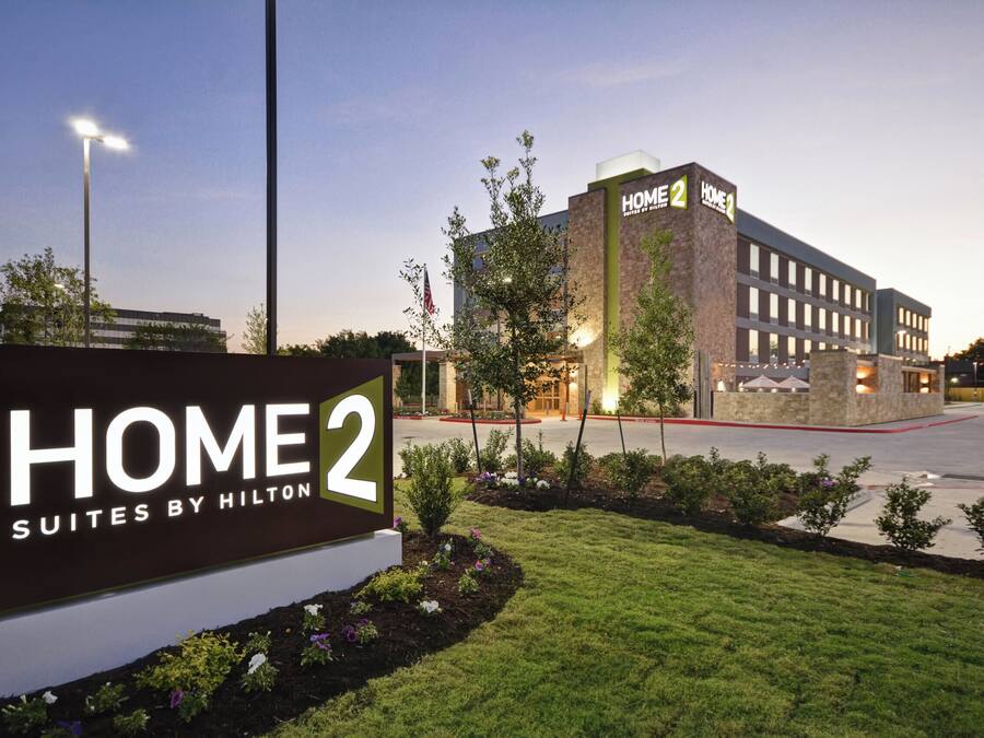 Señal de hotel para Home 2 Suites by Hilton, que muestra un edificio moderno con fachada de piedra y un césped bien cuidado.
