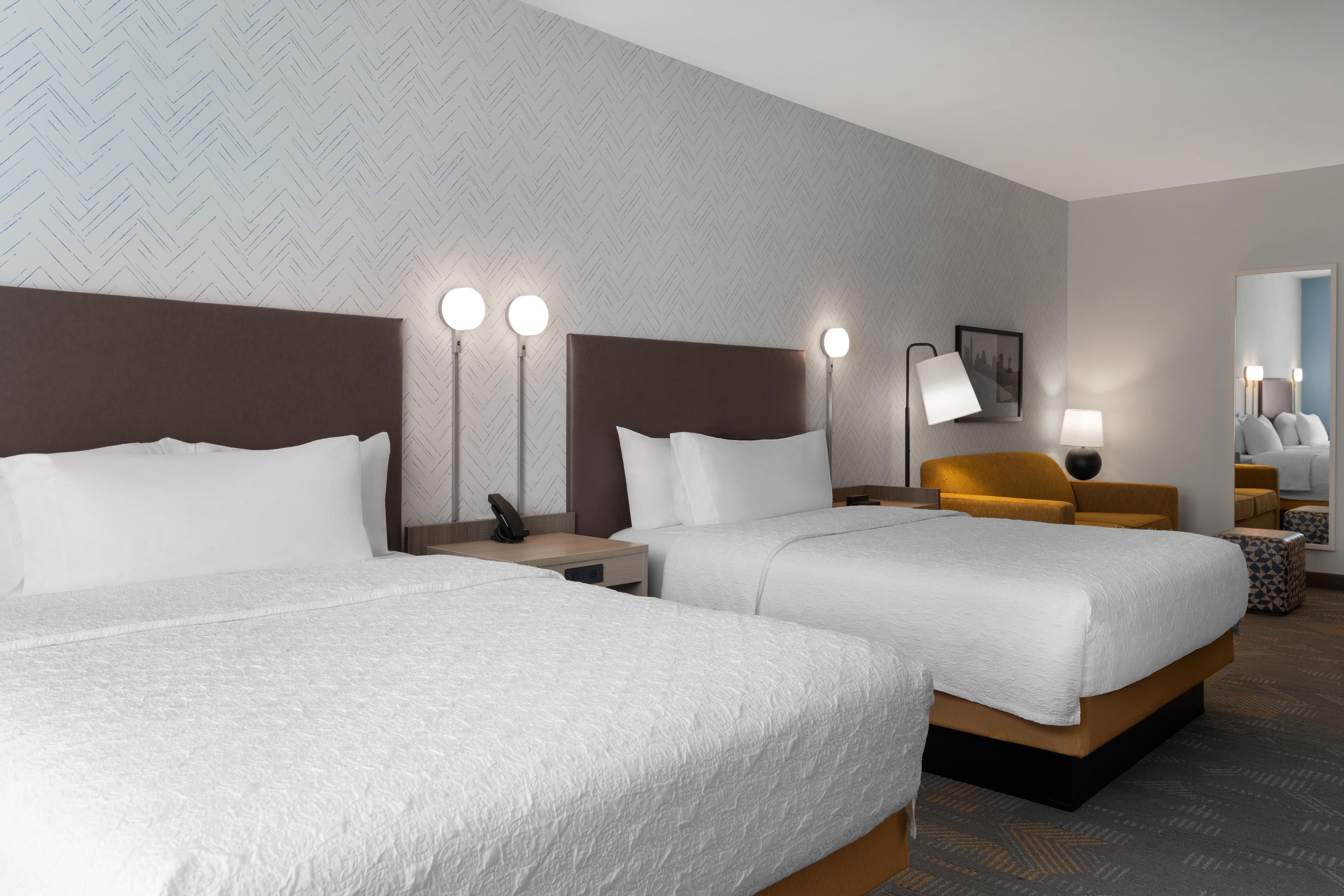 A hotel room with two beds, a nightstand, a lamp, and a mirror.