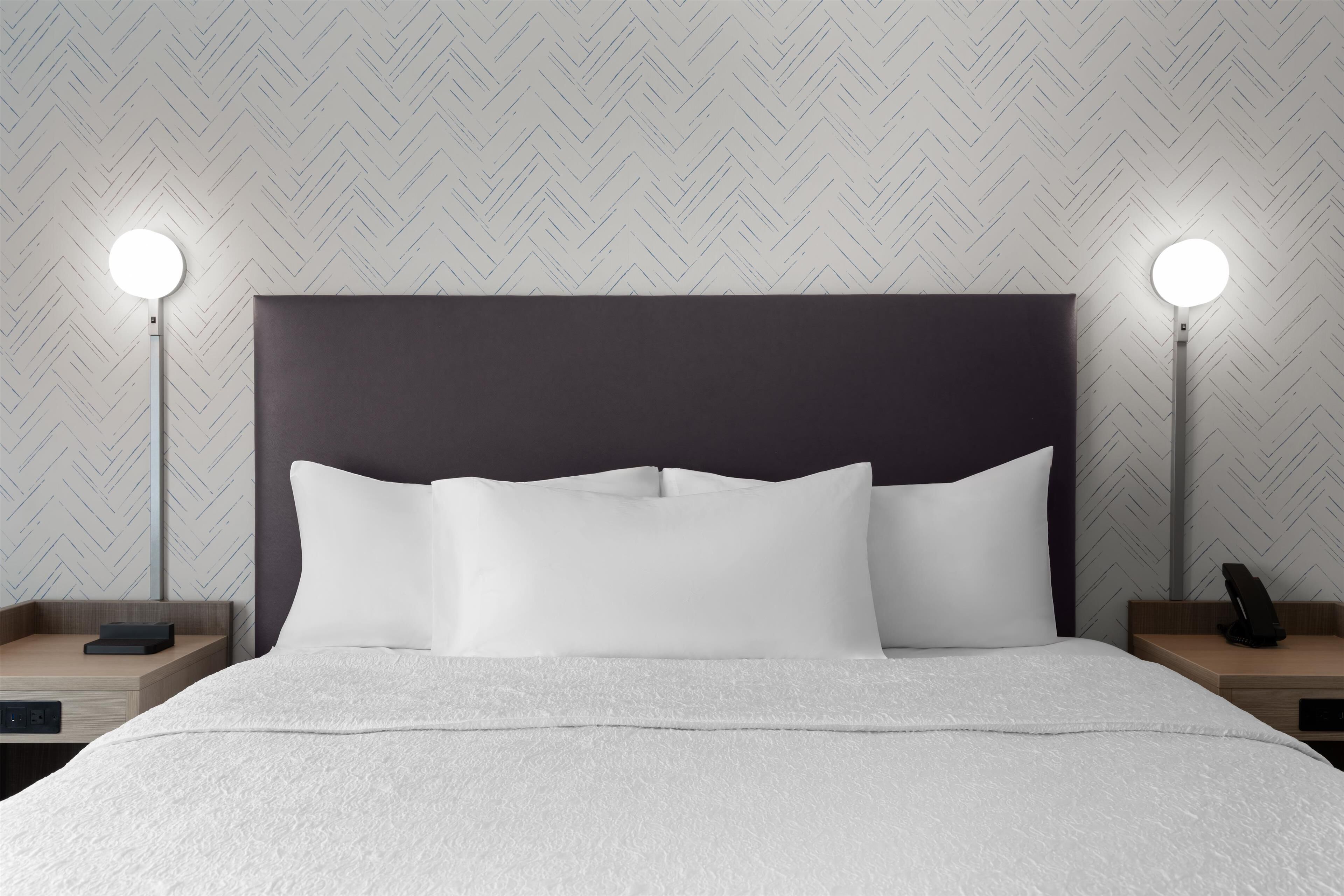 Een net opgemaakt bed met witte lakens, twee nachtkastjes met lampen, en een wand met een herringbone-patroon.