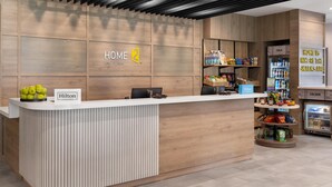 A hotel reception desk with a modern design, featuring a wooden panel wall, a digital display, and a minibar. - Home2 Suites By Hilton Austin South I-35 (Austin)