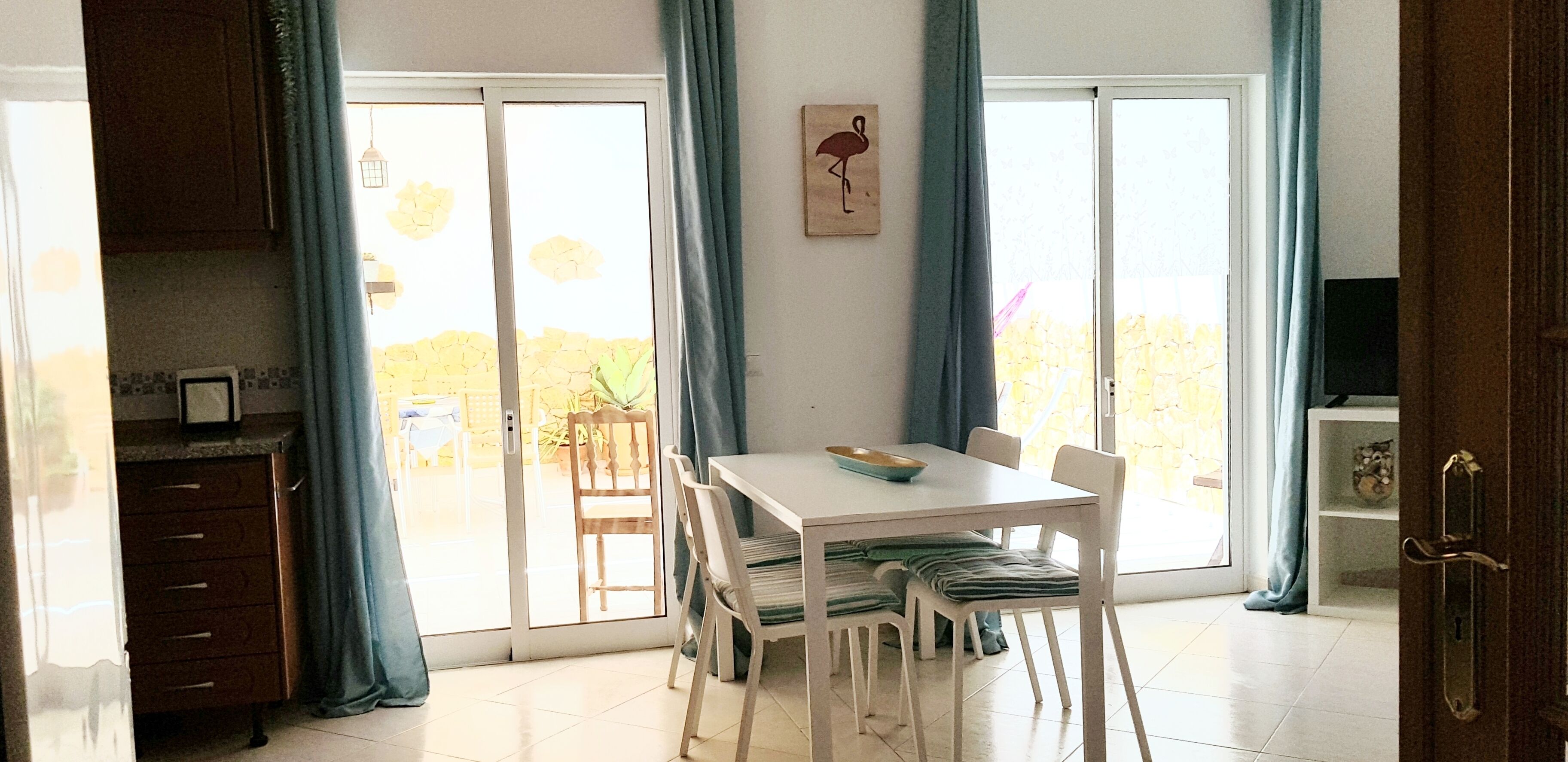 Uma área de refeições com mesa e cadeiras, vista através de uma porta de vidro e área de cozinha com armários e micro-ondas.