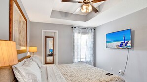 Une chambre à coucher comprenant un lit, un ventilateur de plafond, une télévision et une porte donnant sur une autre pièce.