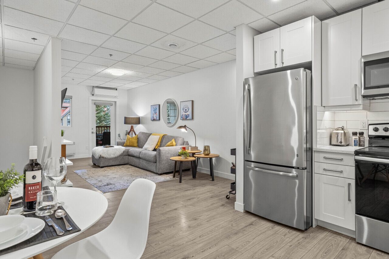 A modern kitchen with stainless steel appliances, a dining area with a white table and chairs, and a living area with a grey sofa and decorative pillows.