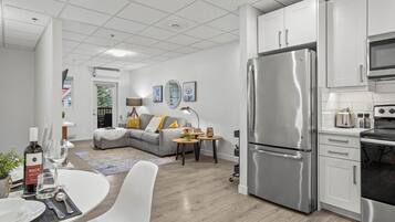 A modern kitchen with stainless steel appliances, a dining area with a white table and chairs, and a living area with a grey sofa and decorative pillows.