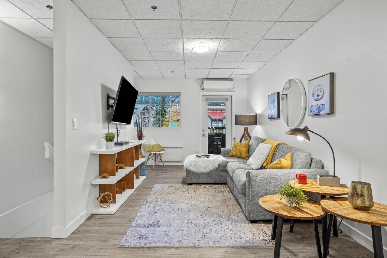 A modern living room with a grey sofa, wooden side tables, a flat-screen TV mounted on the wall, and a large window providing a view of the outside.