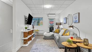 A modern living room with a grey sofa, wooden side tables, a flat-screen TV mounted on the wall, and a large window providing a view of the outside.